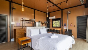 Un dormitorio moderno con cama, mesitas de noche, armario, kitchenette y ventana con vistas a zonas verdes.