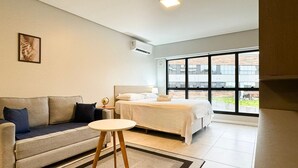 A modern hotel room with a bed, a sofa, a small round table, and a large window offering a view of the outside. - Studio Shopping OKA Floripa - SOK116 (Florianopolis)