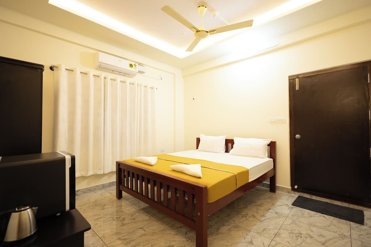Quarto de hotel com cama, ventoinha no teto, ar condicionado, guarda-roupa preto e uma pequena mesa com chaleira.