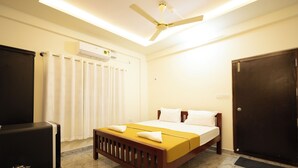 A hotel room with a bed, a ceiling fan, an air conditioning unit, a black wardrobe, and a small table with a kettle. - Golden Crest Residences (Madikeri)