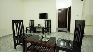 A dining area with a glass-top table, four wooden chairs, a TV mounted on the wall, and a door in the background. - Golden Crest Residences (Madikeri)