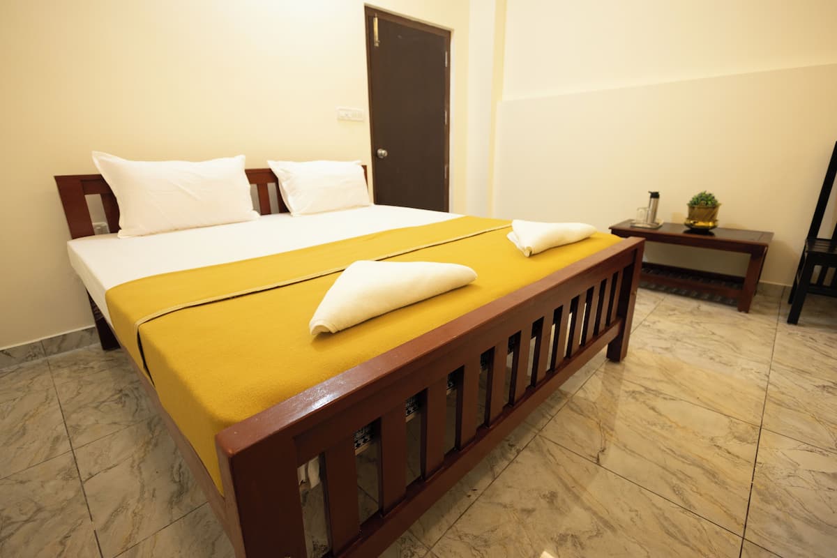 Uma cama bem arrumada com colcha amarela e travesseiros brancos, uma cabeceira de madeira, uma mesinha de cabeceira com uma planta e uma porta ao fundo.