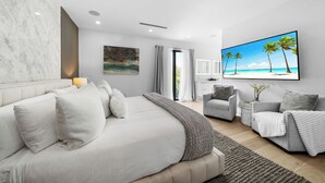 A modern bedroom with a large bed, a grey sofa, a TV, and a painting of a beach scene.
