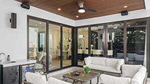 A modern outdoor living area with a glass door leading to a dining area, a small kitchen, and a seating area with a sofa and chairs.