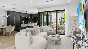 A modern living room with a large white sectional sofa, a dining area with a round table, and a kitchen with dark cabinets and a marble backsplash.