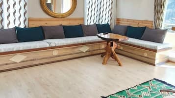Een kamer met een houten vloer, een ronde tafel met houten poten, een bank met kussens, een spiegel en een airconditioning.