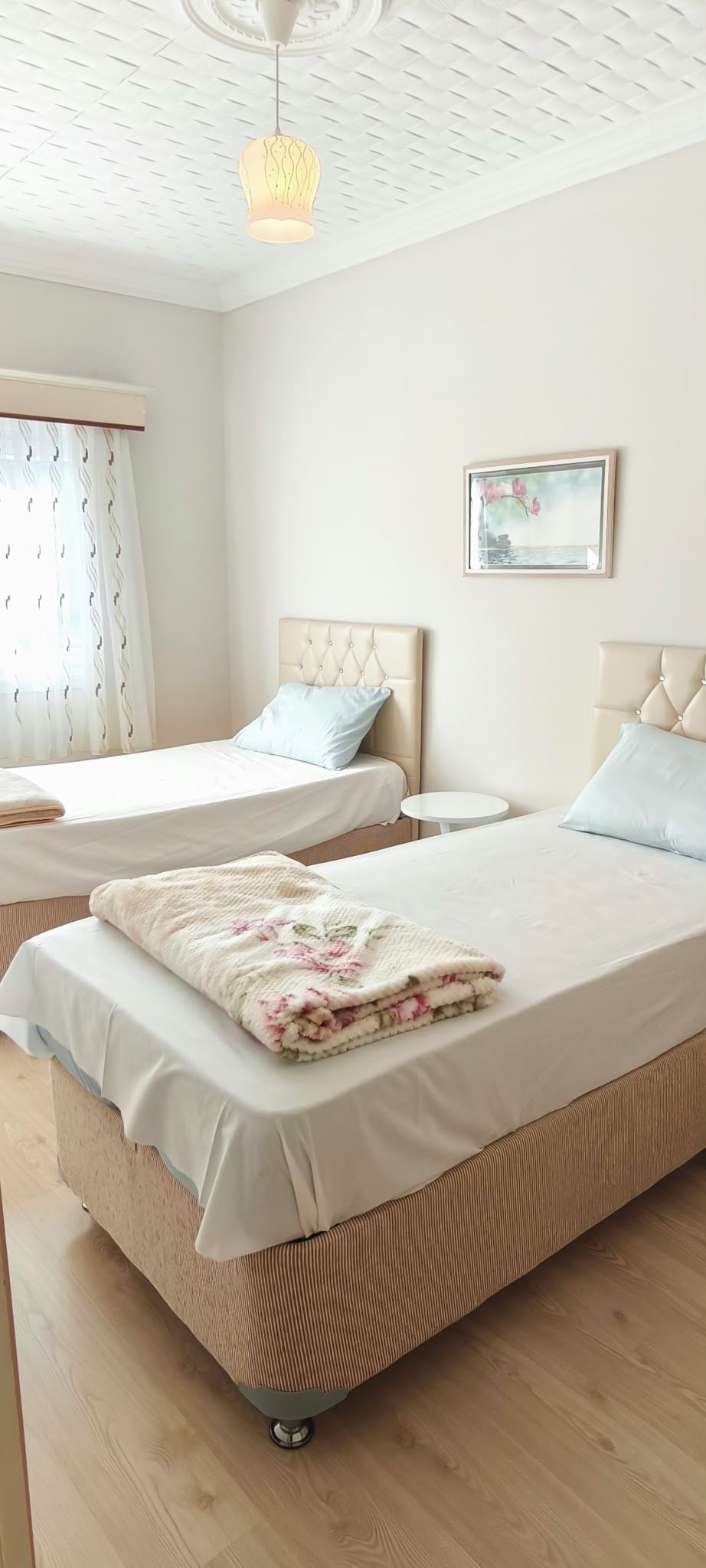 A hotel room with two beds, a round table, a painting on the wall, and a ceiling light fixture.