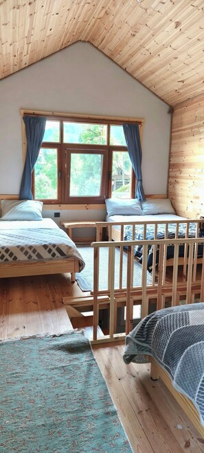 A room with a sloped ceiling, wooden walls, a large window with curtains, two beds, and a view of greenery outside. - YeşilKöy Pansiyon (Çamlıhemşin)