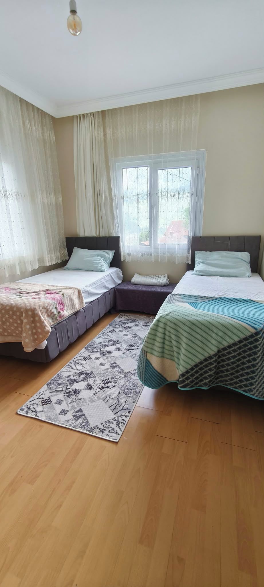 A bedroom with two beds, a window with curtains, a wooden floor, and a light fixture.