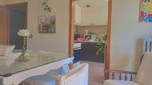 A dining area with a glass table, a white chair, and a vase with a flower. A kitchen is visible in the background with cabinets and appliances. - Elisa Maison (Santa Maria degli Angeli)