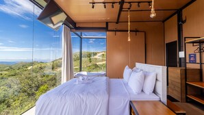 A bedroom with a large bed, a wooden headboard, a bedside table, and a view of a mountainous landscape. - Cabana romântica perto de Florianópolis VST038 (Anitápolis)