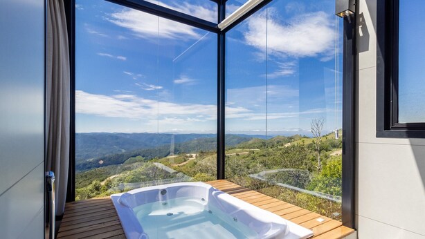 A hot tub on a wooden deck with a glass wall offering a view of a mountainous landscape. - Cabana romântica perto de Florianópolis VST038 (Anitápolis)