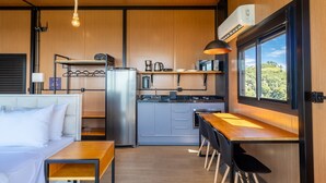 A compact kitchen with a refrigerator, microwave, and coffee maker, a dining area with a table and chairs, and a large window offering a view of greenery. - Cabana romântica perto de Florianópolis VST038 (Anitápolis)