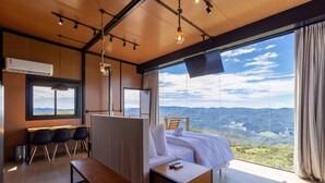A modern hotel room with a large window offering a mountain view, a bed with white linens, a desk with chairs, and a hanging light fixture. - Cabana romântica perto de Florianópolis VST038 (Anitápolis)