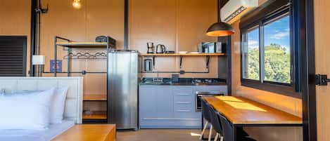 A compact kitchen with a refrigerator, microwave, and coffee maker. There is a dining area with a table and chairs, and a bed with a headboard.