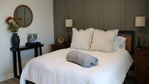 Un lit bien fait, avec du linge de lit blanc, une serviette enroulée et des coussins à rayures. Un miroir rond est accroché au mur, ainsi qu’une petite table sur laquelle est posé un vase de fleurs et deux lampes de chevet.