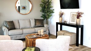 Un salon avec un canapé à carreaux, un miroir rond, une table basse en bois, une table d’appoint avec un vase et un téléviseur fixé au mur.