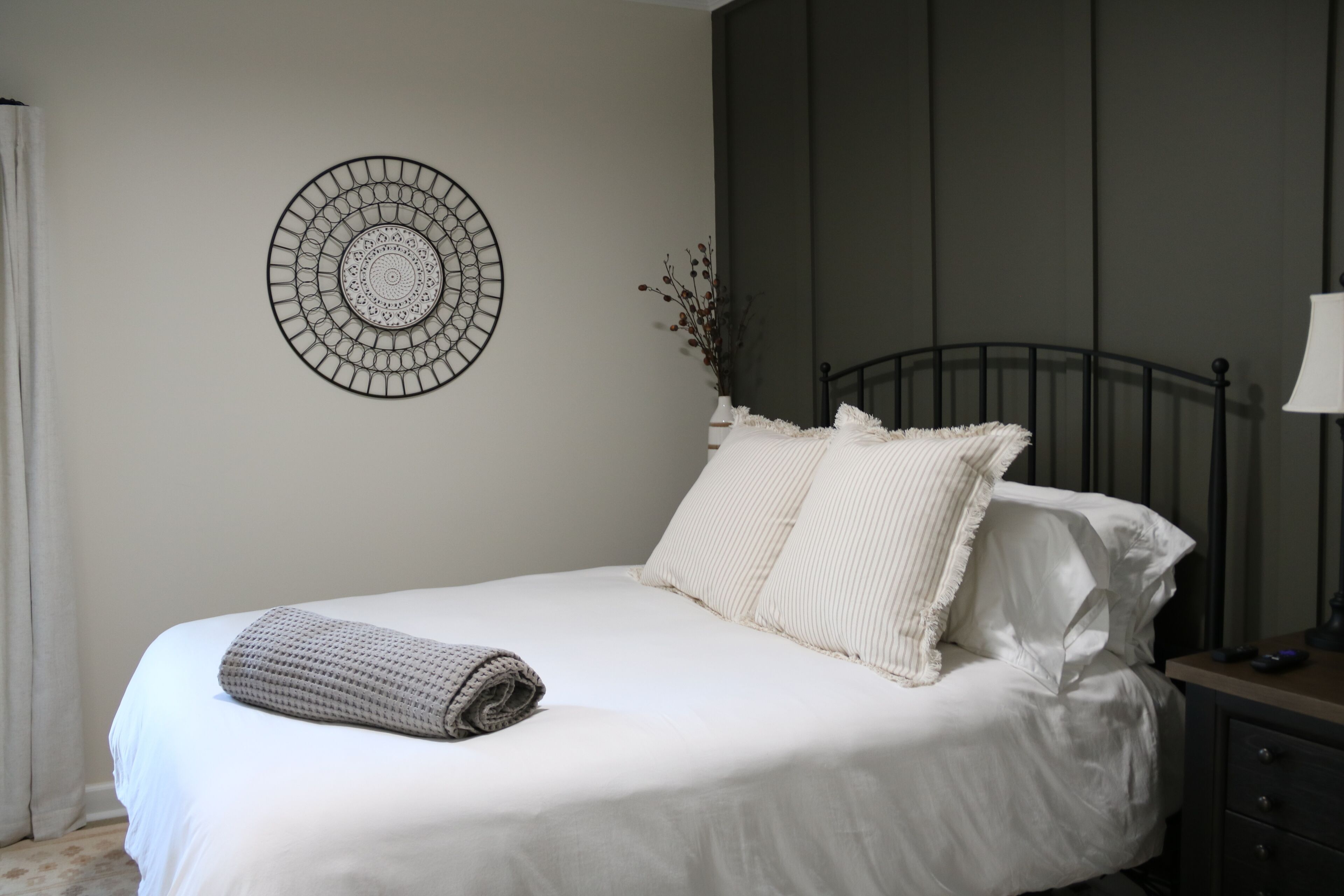 A neatly made bed with white linens and a rolled blanket, a wall-mounted circular decorative piece, and a bedside table with a lamp.