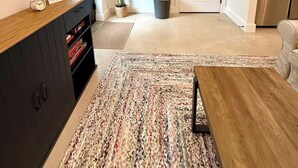 A living room with a flat-screen TV mounted on the wall, a wooden coffee table, a patterned rug, a potted plant, and a door leading to another room.