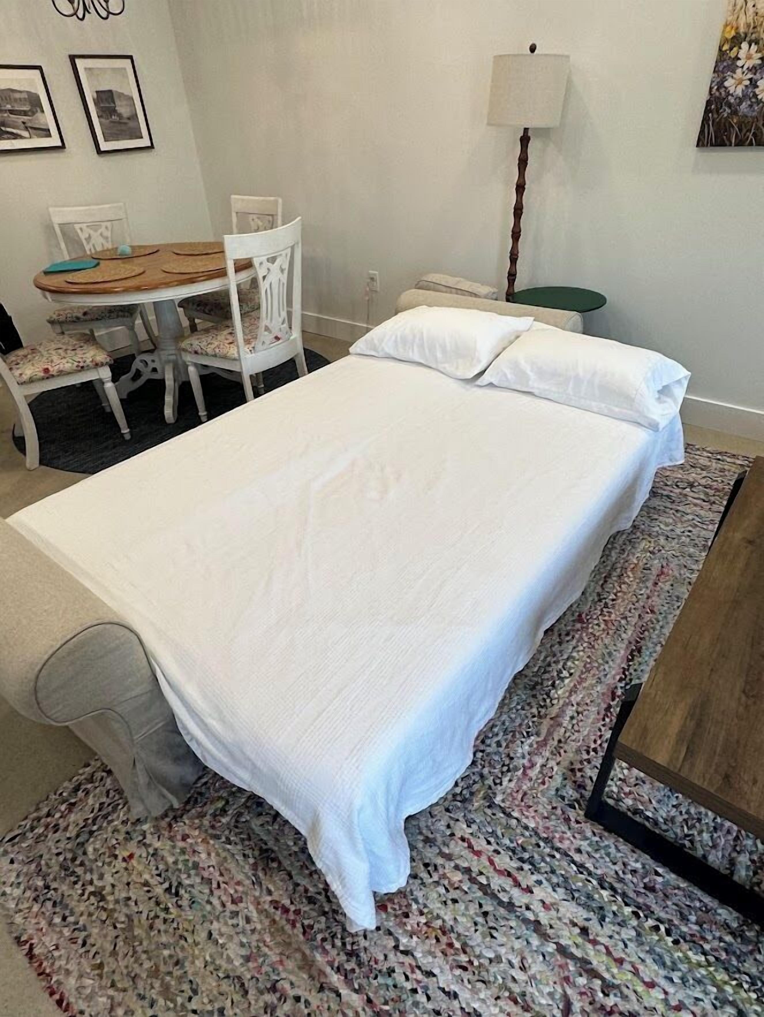 A single bed with white linens, a small round table with chairs, a lamp, and a wall with framed pictures.