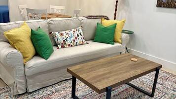 Una sala de estar con sofá, cojines coloridos, una mesa de centro de madera, un tapiz estampado y decoración en la pared.