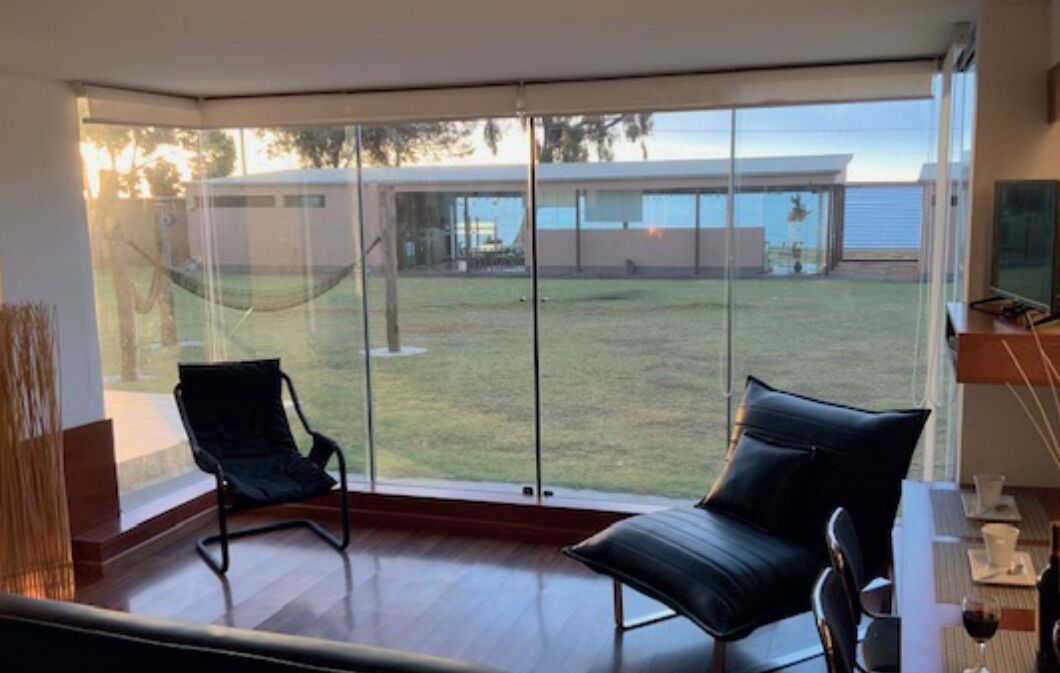 Une chambre moderne dotée d’une grande porte-fenêtre coulissante qui donne sur un espace extérieur verdoyant. On y trouve deux chaises noires et une petite table avec un verre à vin.