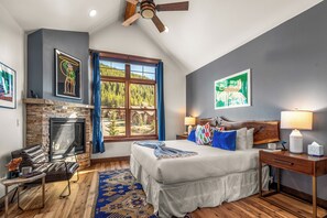 A bedroom with a fireplace, a large window with a view, a bed with pillows, and a nightstand with a lamp. - Modern Mountain Escape - Walk to Lifts - Private Hot Tub, Views, Work-Friendly (Dillon)