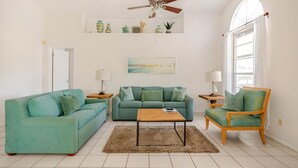 Una sala de estar con un sofá color turquesa, una mesa de centro de madera, un sillón de madera, un ventilador de techo, una ventana con cortinas y una estantería con adornos.