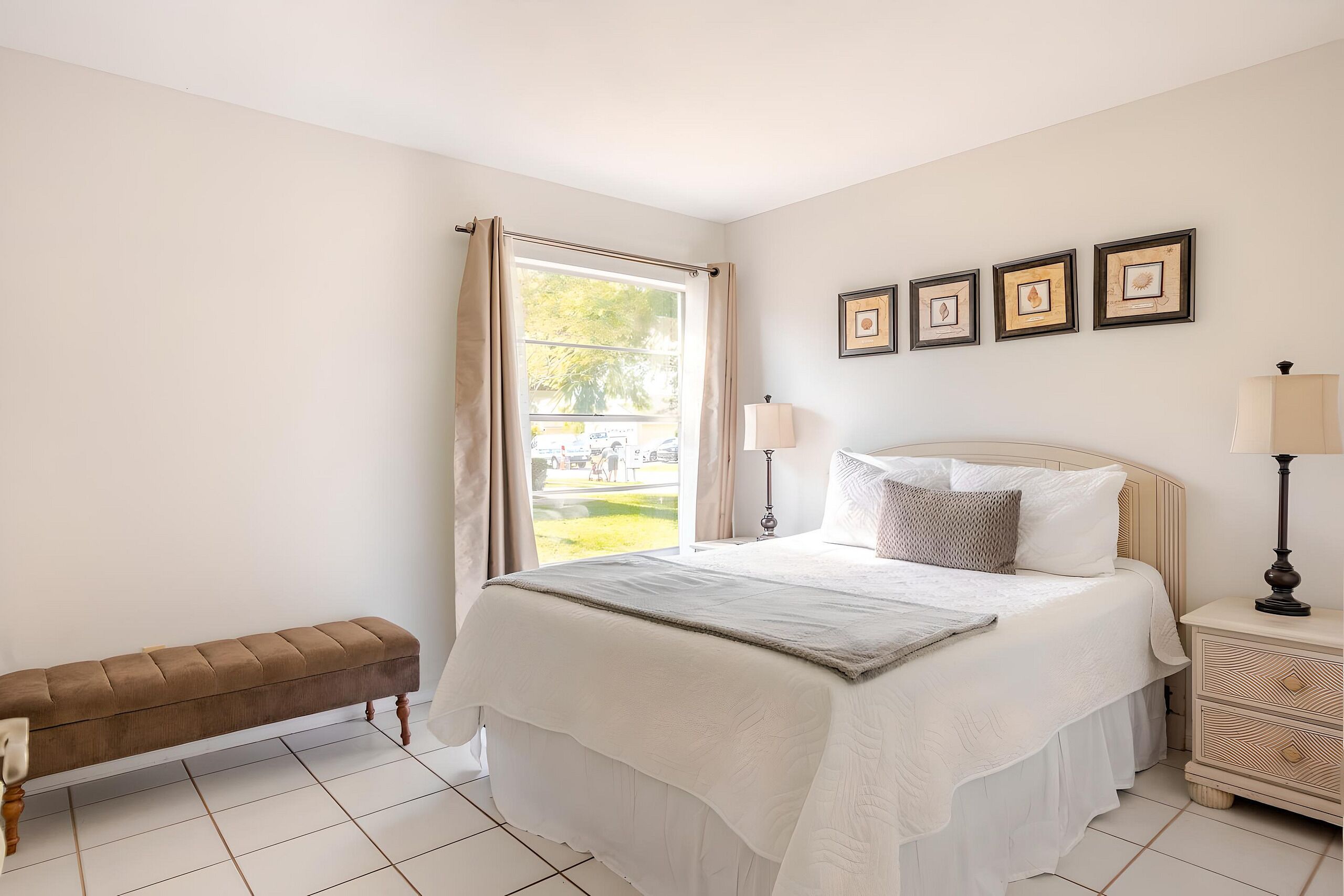A bedroom with a bed, bench, bedside table, lamp, and framed pictures on the wall.