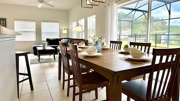 A dining area with a wooden table set for six, surrounded by chairs. There is a ceiling fan, large windows with blinds, and a view of greenery outside.