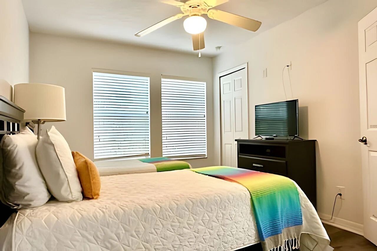 A bedroom with a bed, a ceiling fan, a television on a stand, and a window with blinds.
