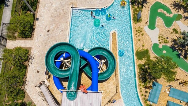 Vue aérienne d’un parc aquatique avec une grande piscine, plusieurs toboggans et un espace de jeux verdoyant.