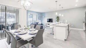 A modern kitchen with a central island, a dining area with a white table set for four, and a living area with a blue sofa. - 4BR Townhome | Private Pool | 10 min to Disney (Kissimmee)
