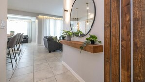 A modern living room with a grey sofa, a wooden shelf with plants, a large mirror, and a dining area with chairs and a table. - 2BR Storey Lake | Sleeps 7 | 10min Disney (Kissimmee)