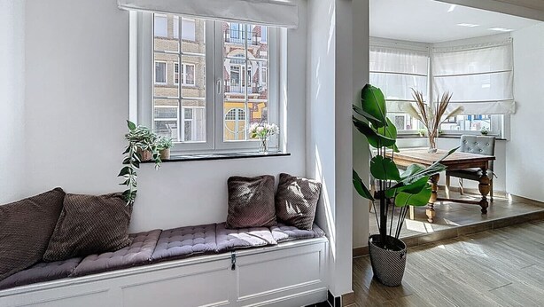 A modern living room with a built-in bench, a large window with a view of buildings, a dining area with a table and chairs, and a potted plant. - Maison 't Blyhuis - De Panne (De Panne)