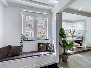 Ein modernes Wohnzimmer mit einer eingebauten Bank, einem großen Fenster mit Blick auf Gebäude, einem Essbereich mit Tisch und Stühlen sowie einer Zimmerpflanze.