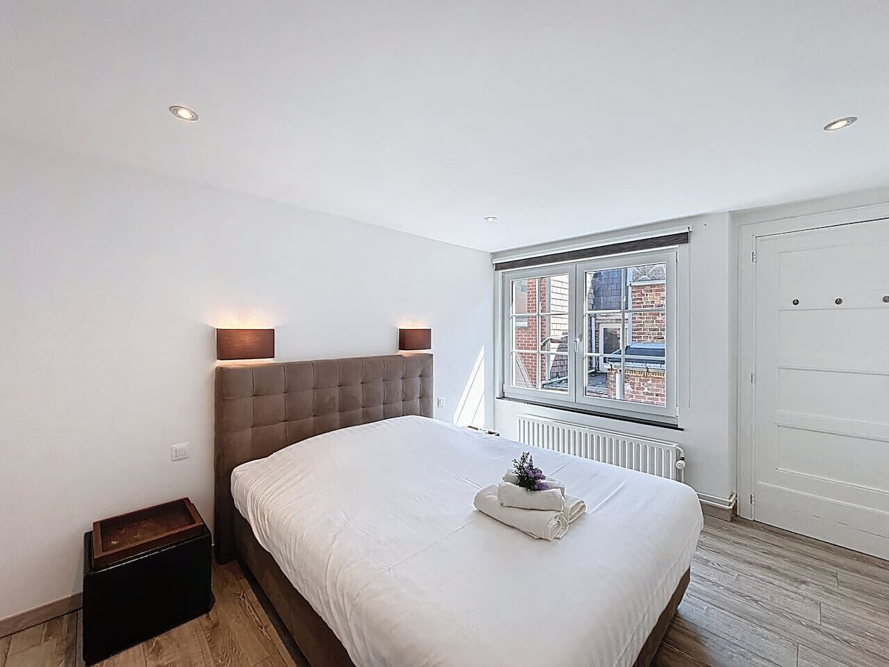 Een slaapkamer met een groot bed, een nachtkastje, een raam met uitzicht op gebouwen en een radiator.