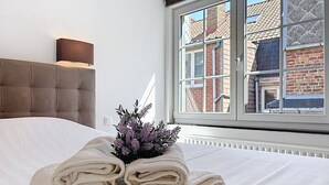 A neatly made bed with rolled towels and a small plant, a window with a view of a brick building, and a wall-mounted lamp. - Maison 't Blyhuis - De Panne (De Panne)