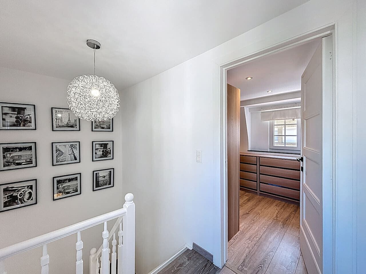 A hallway with a staircase, a chandelier, and a series of framed photographs on the wall.