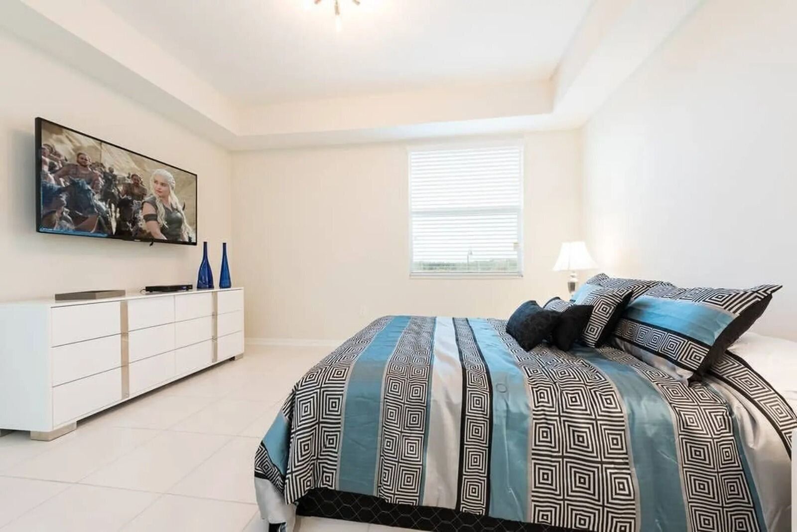 A bedroom with a bed, a TV on the wall, a window, and a bedside table with a lamp.