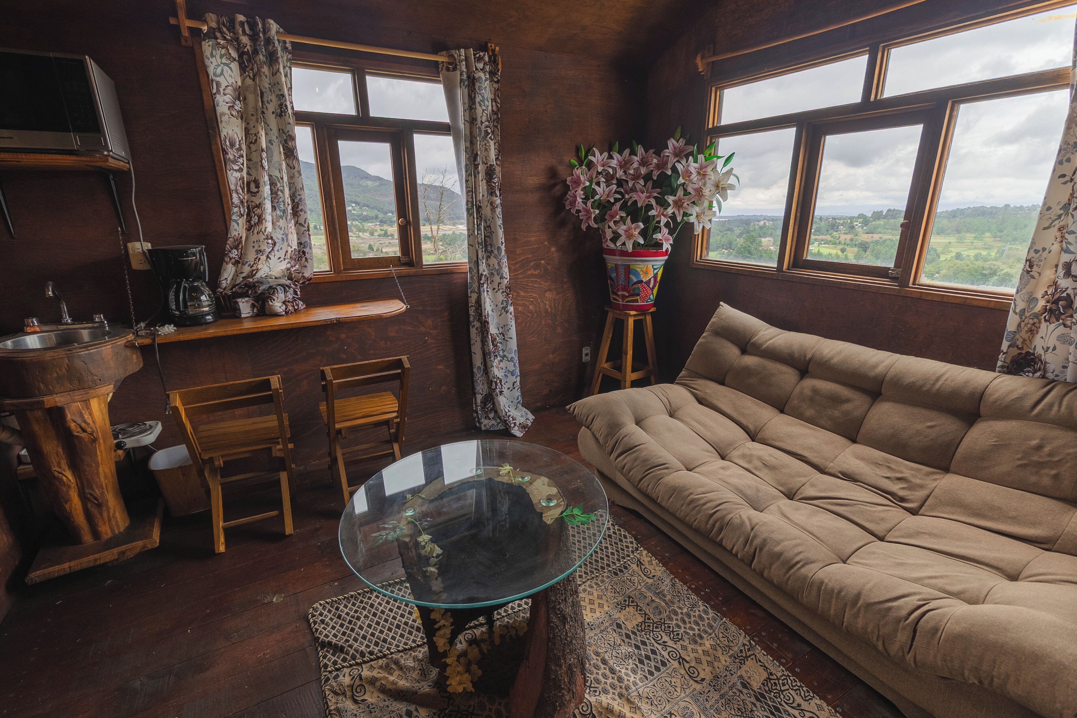 Una sala de estar acogedora con un sofá, una mesa de centro de vidrio, sillas de madera, una mesa de comedor de madera y vistas al campo a través de ventanas grandes.
