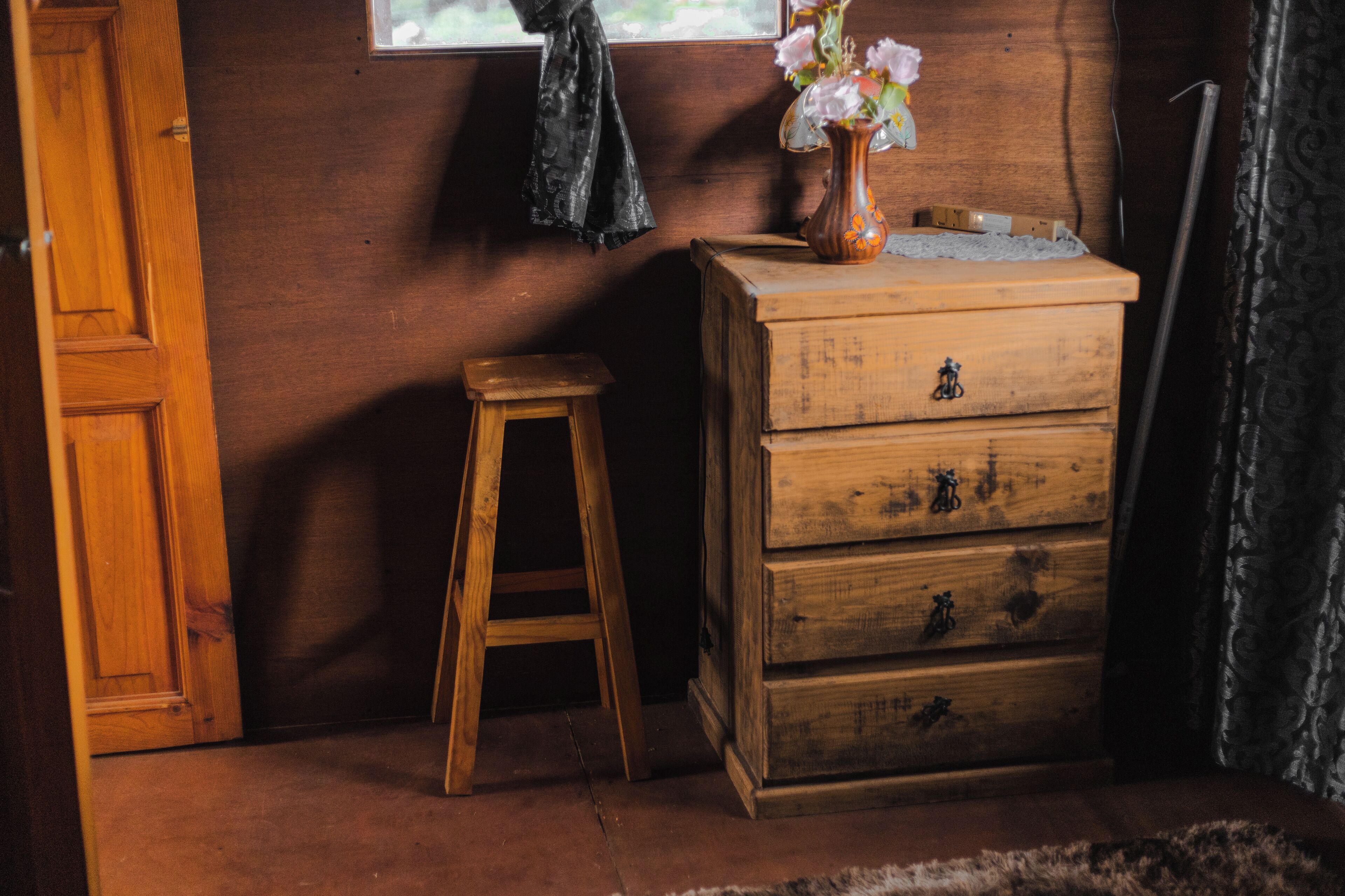 A wooden dresser with drawers, a small stool, a window with a curtain, and a vase with flowers on the dresser.