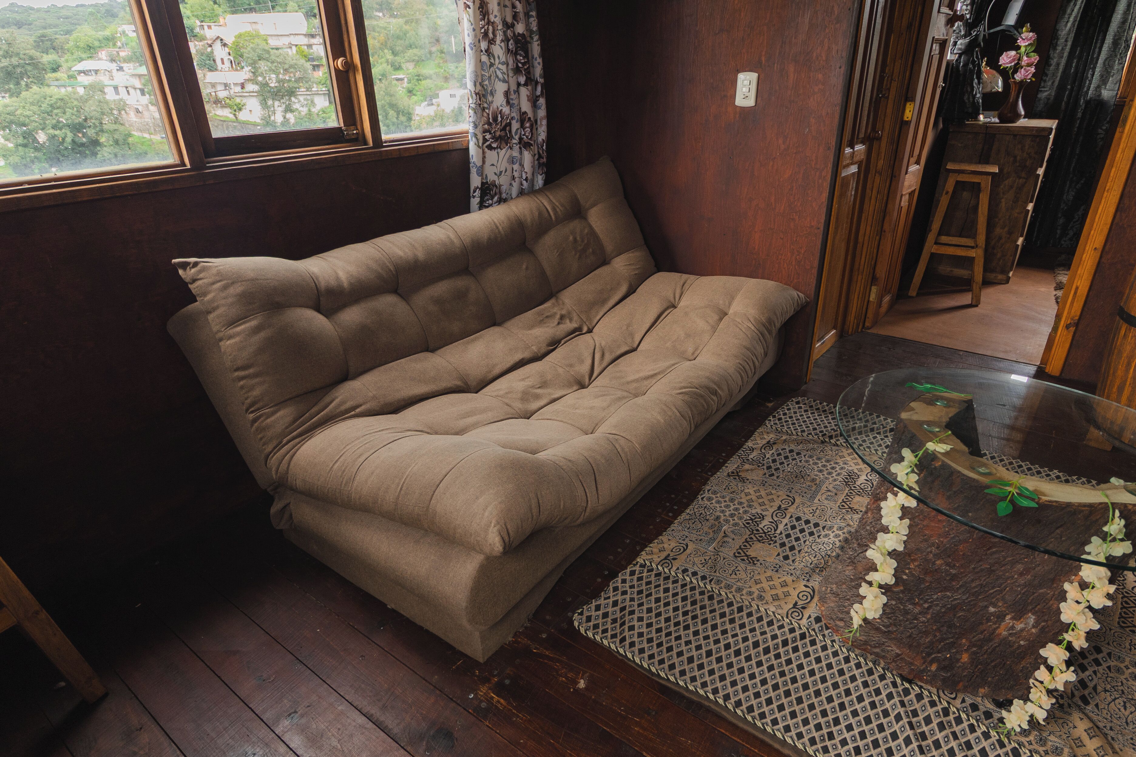 Una habitación con sofá, una mesa de centro de vidrio, un tapete estampado y vistas a través de una ventana.
