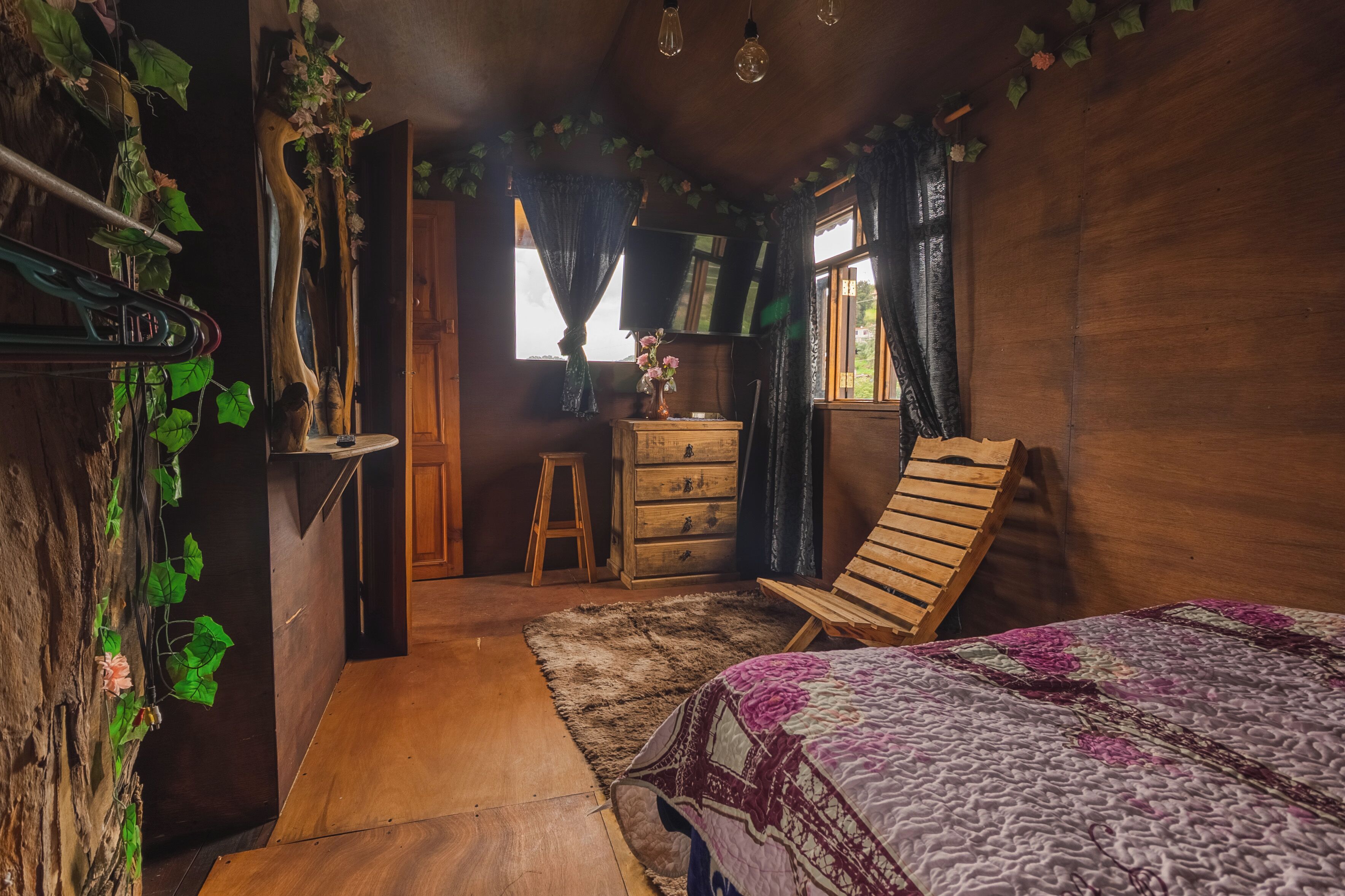 Una habitación con una cama de madera, un aparador de madera, un taburete pequeño y una ventana con cortinas.
