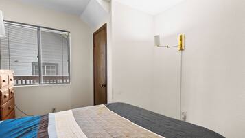 Une chambre à coucher comprenant un lit, une commode, une fenêtre avec des stores et un luminaire fixé au mur.