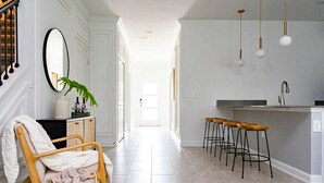 Un pasillo luminoso que conduce a una cocina con barra, una silla de mimbre y un espejo en la pared.