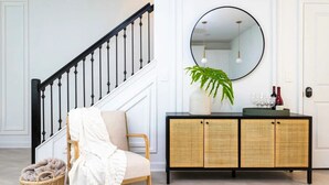 Una escalera con balaustres negros, una silla de mimbre con una manta blanca, una consola de mimbre con un espejo y una planta en maceta.