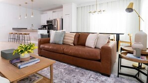 Una sala moderna con un sofá de cuero marrón, una mesa de centro de madera con libros, una pequeña planta en maceta y un comedor con una mesa y sillas blancas.