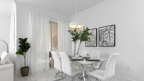 A modern dining area with a white table set for four, surrounded by white chairs. There are two wall-mounted artworks above the table, a potted plant on the table, and a large window with white curtains. - Storey Lake 4BR Townhome w/ Private Pool (Kissimmee)