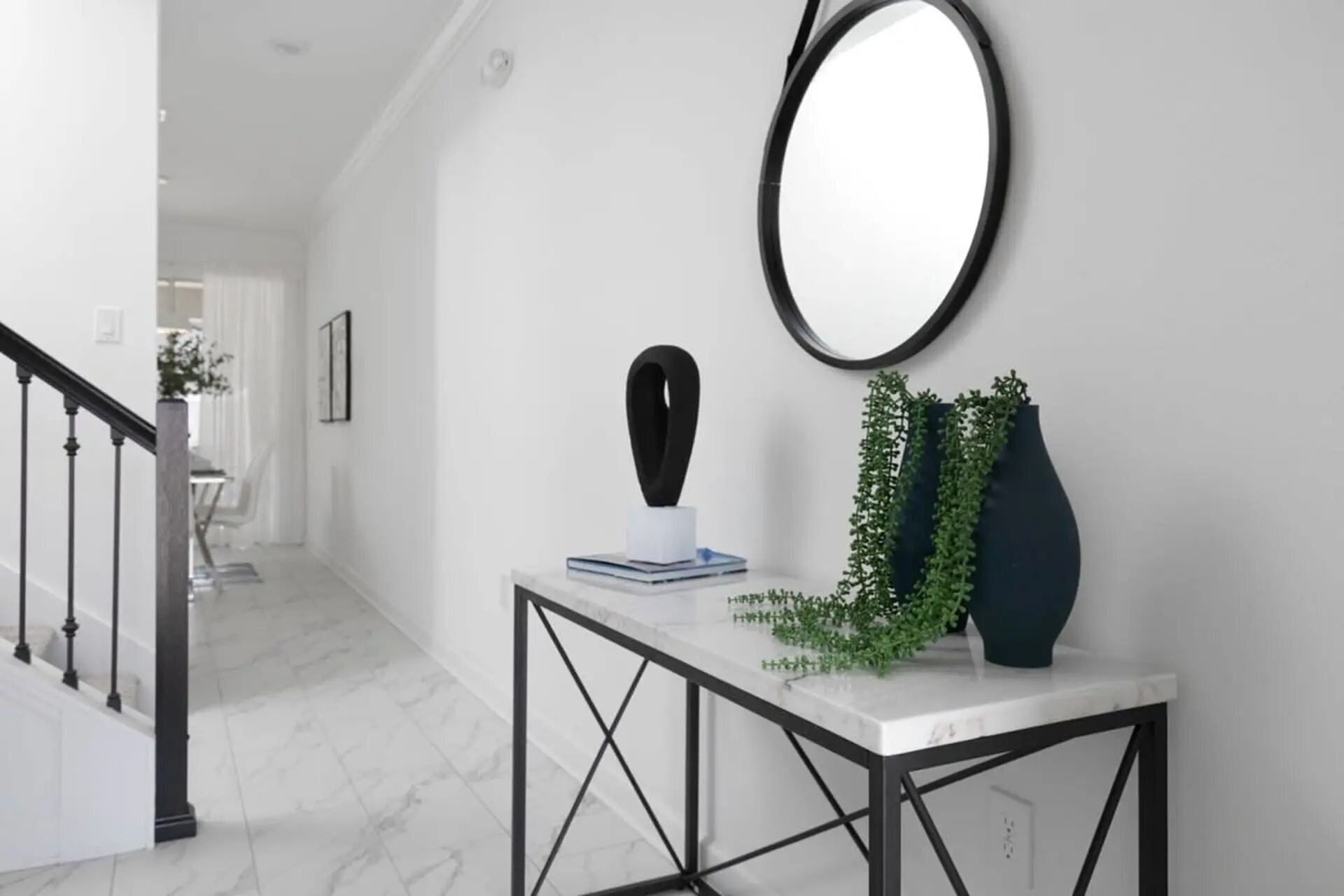 Un pasillo con suelo de mármol blanco, una consola de metal negro con un jarrón blanco y una escultura negra, y un espejo redondo en la pared.